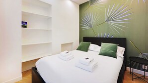 A neatly made bed with white linens and two green pillows, a wall with a decorative leaf pattern, and a shelf unit with a book titled "Italy". - Serene Apartment - 1br/4p - Place de la Bastille (Paris)