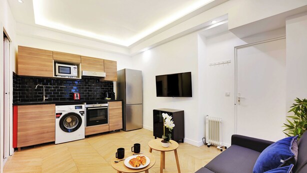 A compact living space with a kitchenette, a sofa, a small table with a plate of food, and a potted plant. - Serene Apartment - 1br/4p - Place de la Bastille (Paris)