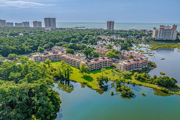 An aerial view of a residential complex with multiple buildings, a large body of water, and surrounding greenery.
