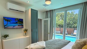 Ein Schlafzimmer mit einem Bett, einem Fernsehgerät an der Wand, einem Fenster mit Blick auf einen Swimmingpool und einer Klimaanlage.
