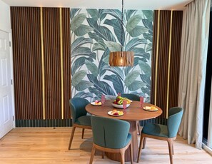 Un coin repas avec une table ronde en bois, quatre chaises à l’assise rembourrée de couleur verte, un saladier rempli de fruits et un luminaire suspendu.