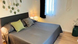 A neatly made bed with a grey quilt, two pillows, and a green accent pillow. There is a bedside lamp, a wall hanging with leaves, and a green vase with white flowers on the floor.