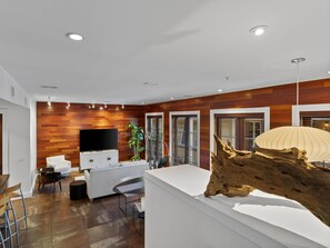 A modern living room with a wooden wall, a flat-screen TV, a white sofa, a dining table, and a large piece of driftwood.