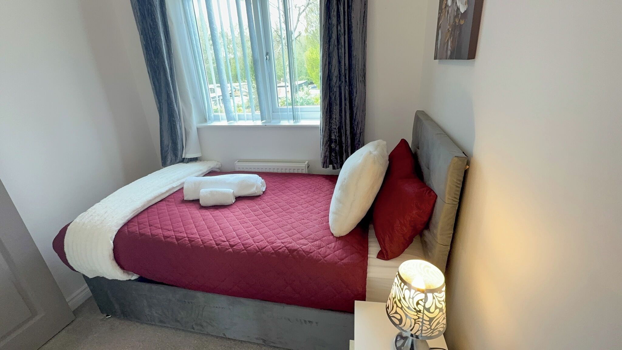Un dormitorio con cama, almohadas, una lámpara de noche y una ventana con cortinas.