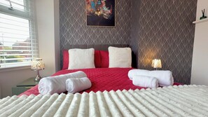 A neatly made bed with a red quilt, white pillows, and a white fluffy blanket. There are two bedside lamps and a painting on the wall. - Manchester Home Away From Home (Manchester)