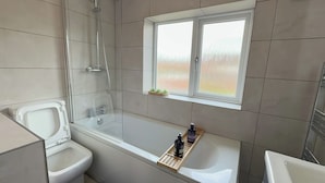 A modern bathroom with a bathtub, toilet, and shower. There is a window above the bathtub with a view of trees. - Manchester Home Away From Home (Manchester)