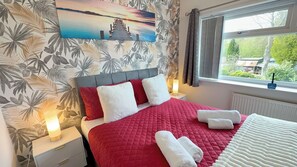 A bedroom with a red quilted bedspread, white pillows, and a painting of a sunset over a pier. - Manchester Home Away From Home (Manchester)