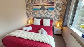A hotel room with a red bed, white pillows, a tropical wallpaper, a sunset painting, and a window with a view. - Manchester Home Away From Home (Manchester)