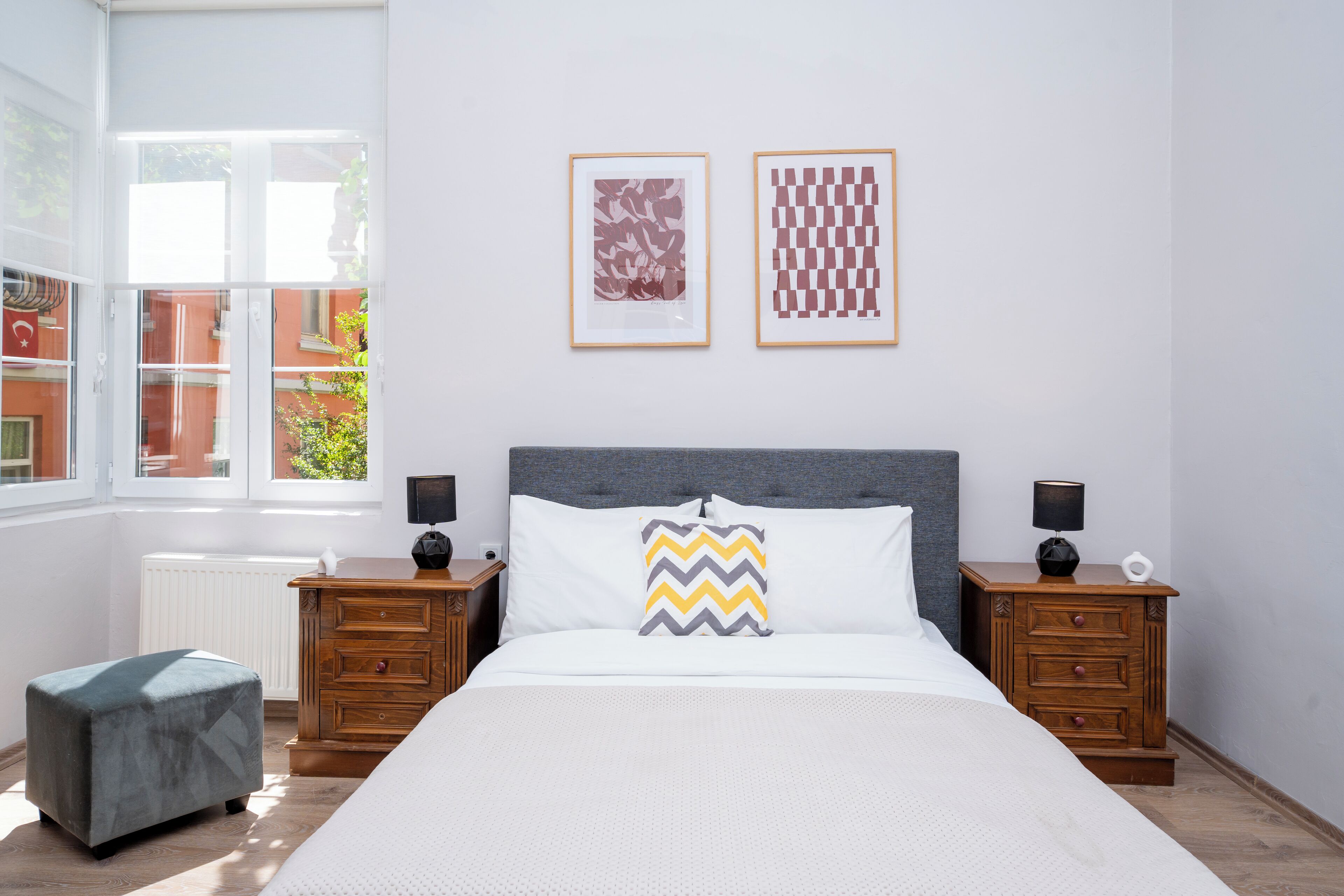 A bedroom with a large bed, two bedside tables, a nightstand, a window, and two framed pictures on the wall.