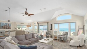 A spacious living room with a large gray sectional sofa, a white coffee table, and striped armchairs. A ceiling fan and pendant lights are visible. - 800 Luxury 8BR Oceanfront Retreat w/Pool & Hot Tub (Nags Head)