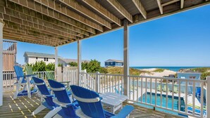 A covered deck with blue Adirondack chairs, a pool, and a view of the beach and ocean. - 800 Luxury 8BR Oceanfront Retreat w/Pool & Hot Tub (Nags Head)