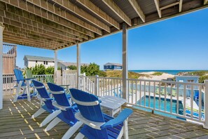 A covered deck with blue Adirondack chairs, a pool, and a view of the beach and ocean.