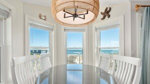 A dining area with a glass table, white chairs, a wicker ceiling light, and ocean views through large windows. - 800 Luxury 8BR Oceanfront Retreat w/Pool & Hot Tub (Nags Head)