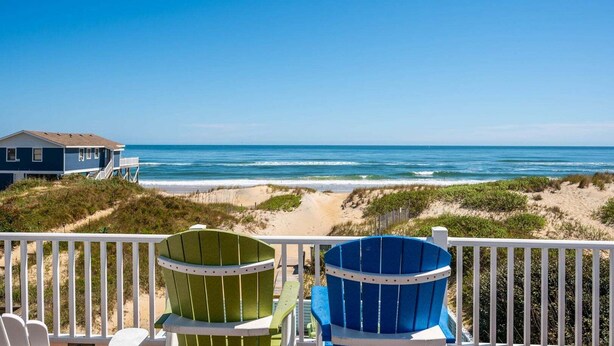 A beach view from a balcony with two Adirondack chairs, a house, and a sandy beach. - 800 Luxury 8BR Oceanfront Retreat w/Pool & Hot Tub (Nags Head)