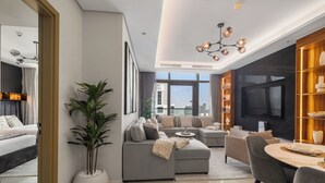 A modern living room with a grey sofa, a dining table, and a large TV. There is a large window with a city view and a potted plant. - Gogoto LUX Burj View Spacious (Dubai)