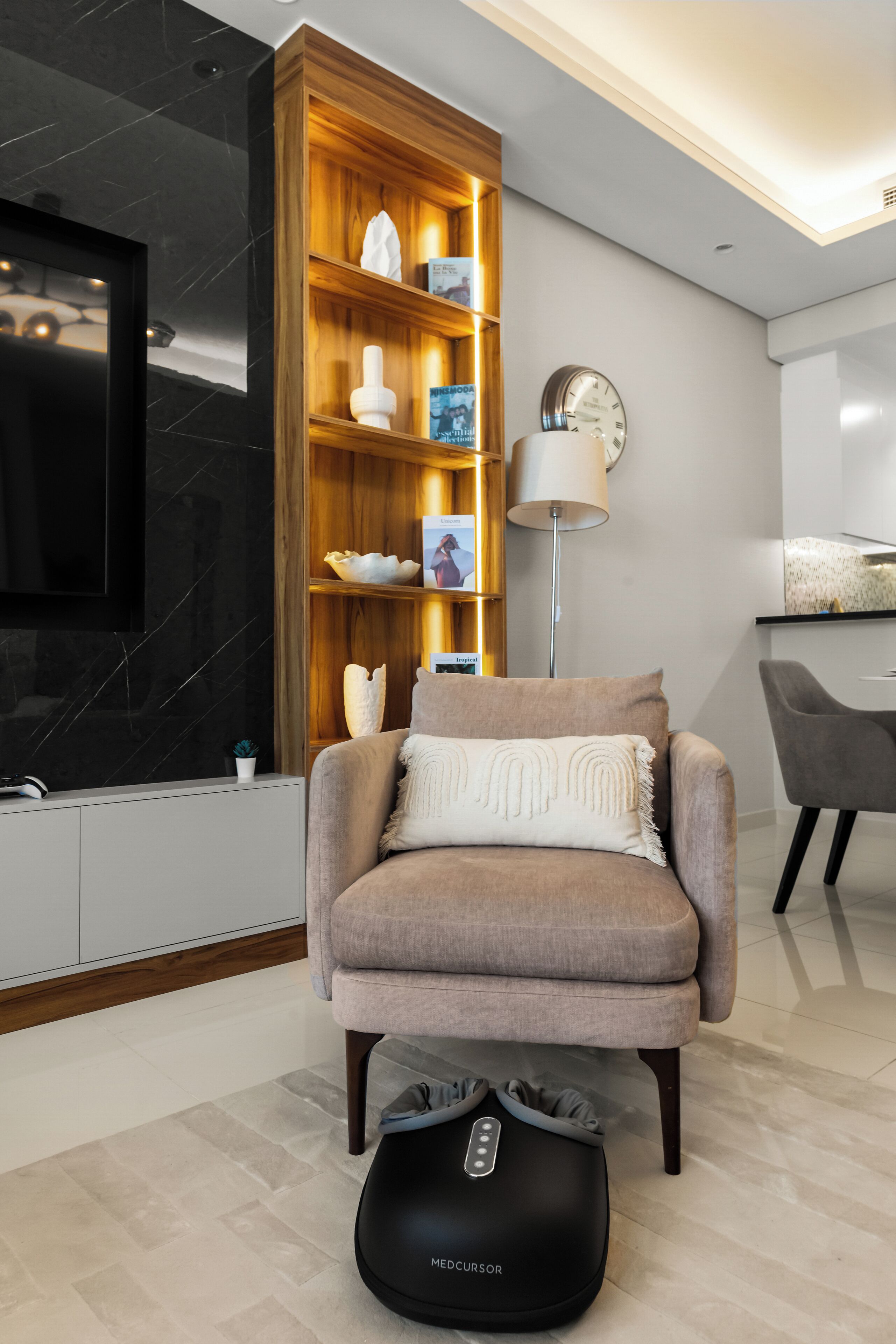 A modern living room with a sofa, a massage chair, a massage device, a wooden shelving unit with decorative items, a wall clock, and a flat-screen TV.