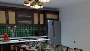 Un coin repas avec une table en bois dressée pour quatre personnes, entourée de chaises, une cuisine en arrière-plan et des lampes suspendues.