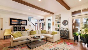 Ein geräumiges Wohnzimmer mit einer großen Couch, einem Couchtisch und einem farbenfrohen Teppich. Ein Fernseher ist an der Wand befestigt, eine Treppe führt zu einer oberen Ebene, und eine Tür ist mit einem Spiegel und Dekorationsgegenständen versehen.