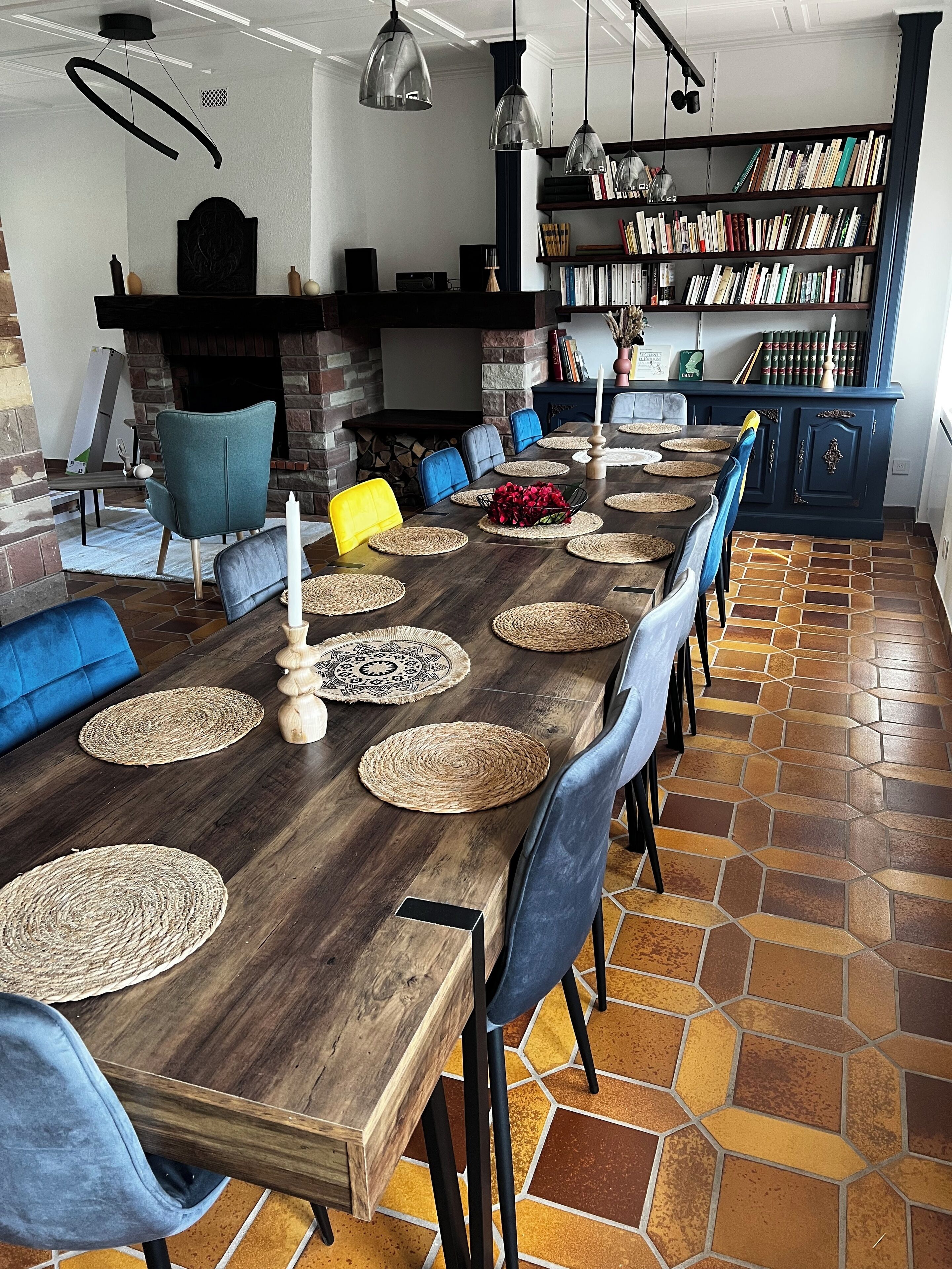 A dining area with a long wooden table, blue and yellow chairs, woven placemats, and a bookshelf filled with books.