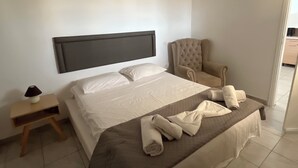 Habitación de hotel con una cama grande, una mesita de noche con lámpara, una silla y vistas al pasillo.