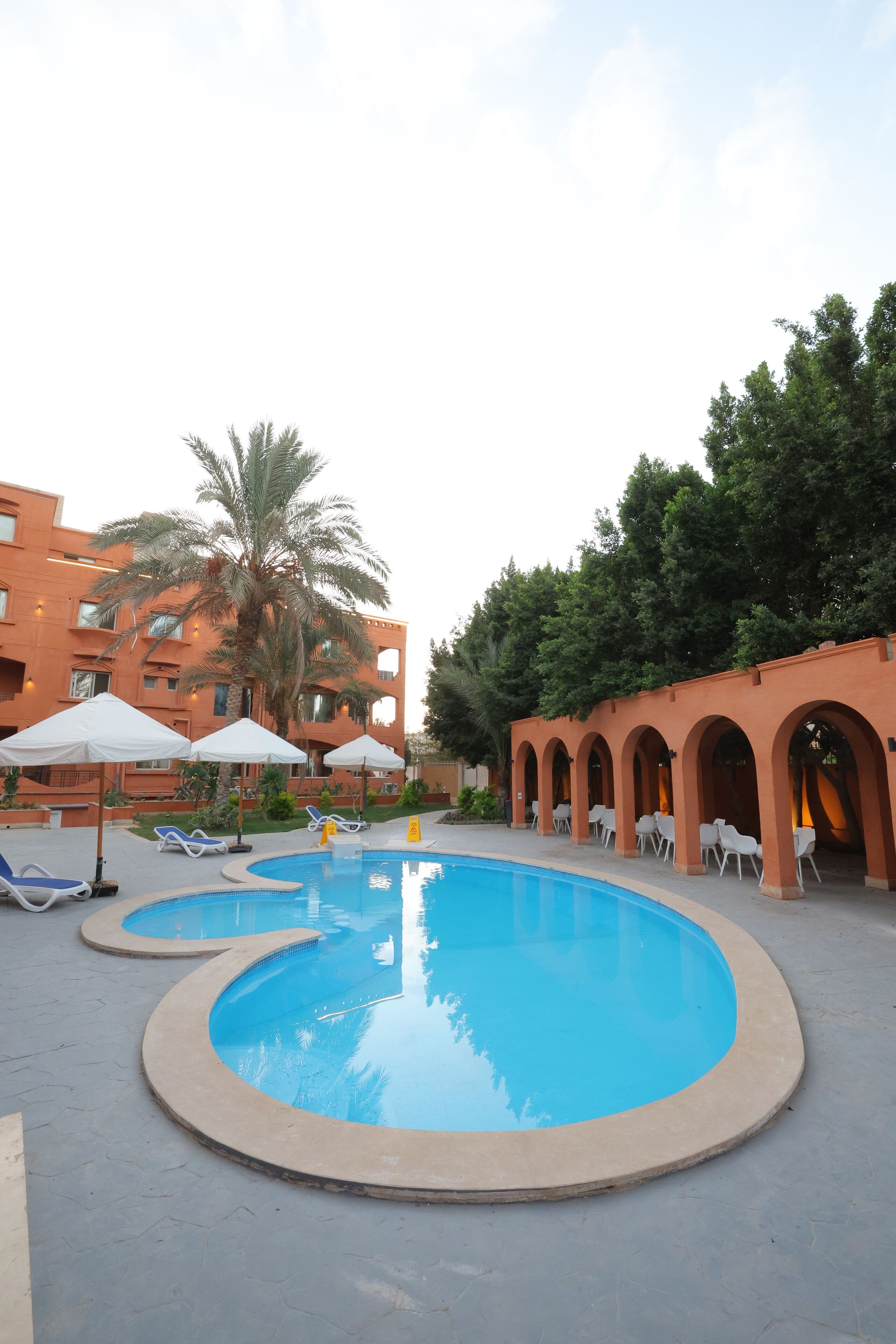Una piscina con sillas de descanso y sombrillas, rodeada de un edificio con arcos y palmeras.
