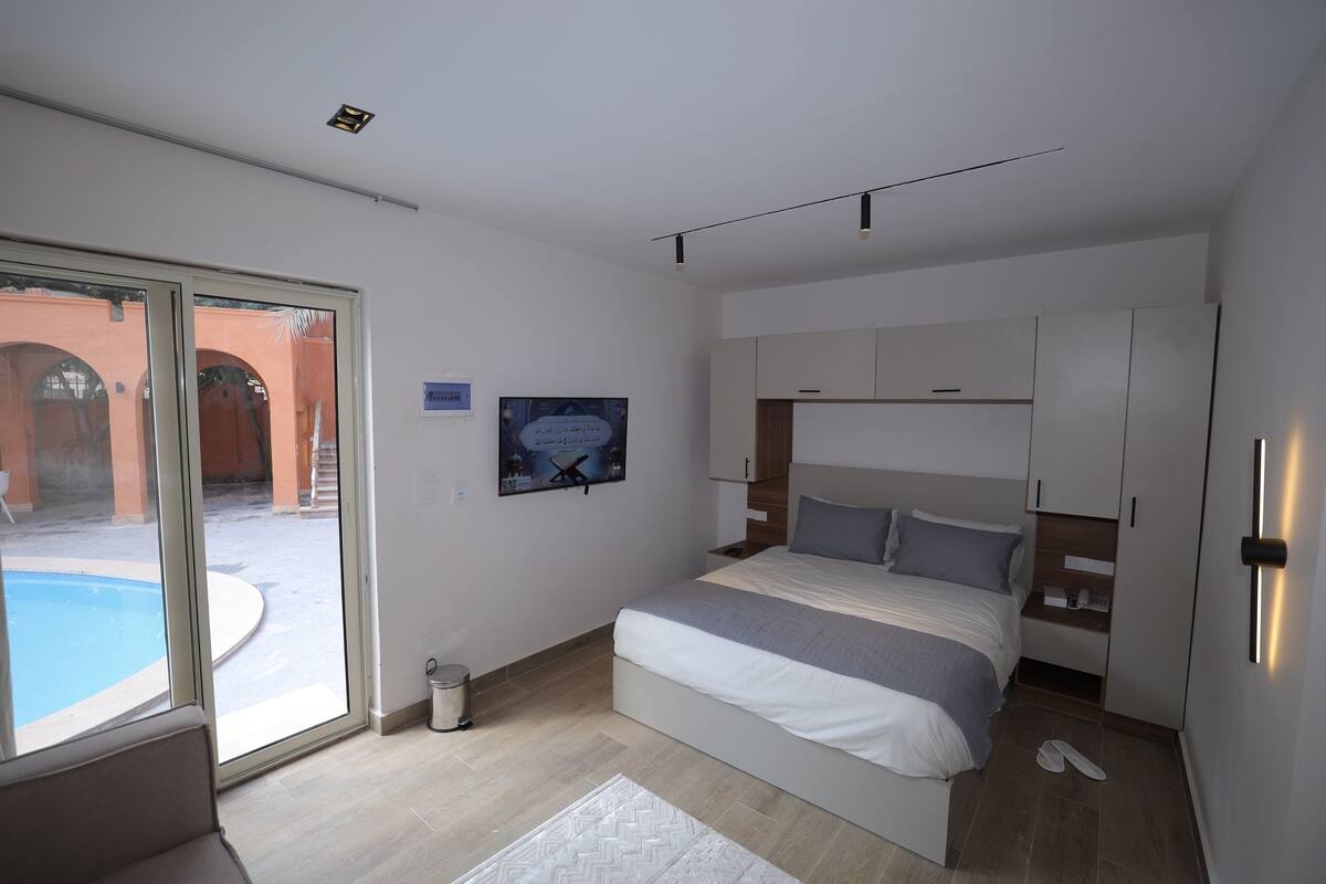 A modern bedroom with a large bed, built-in wardrobe, and a sliding glass door leading to a pool area.