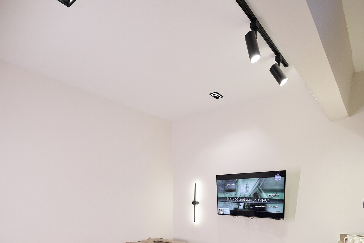 A modern living room with a sofa, a coffee table, a TV mounted on the wall, and a kitchen area.