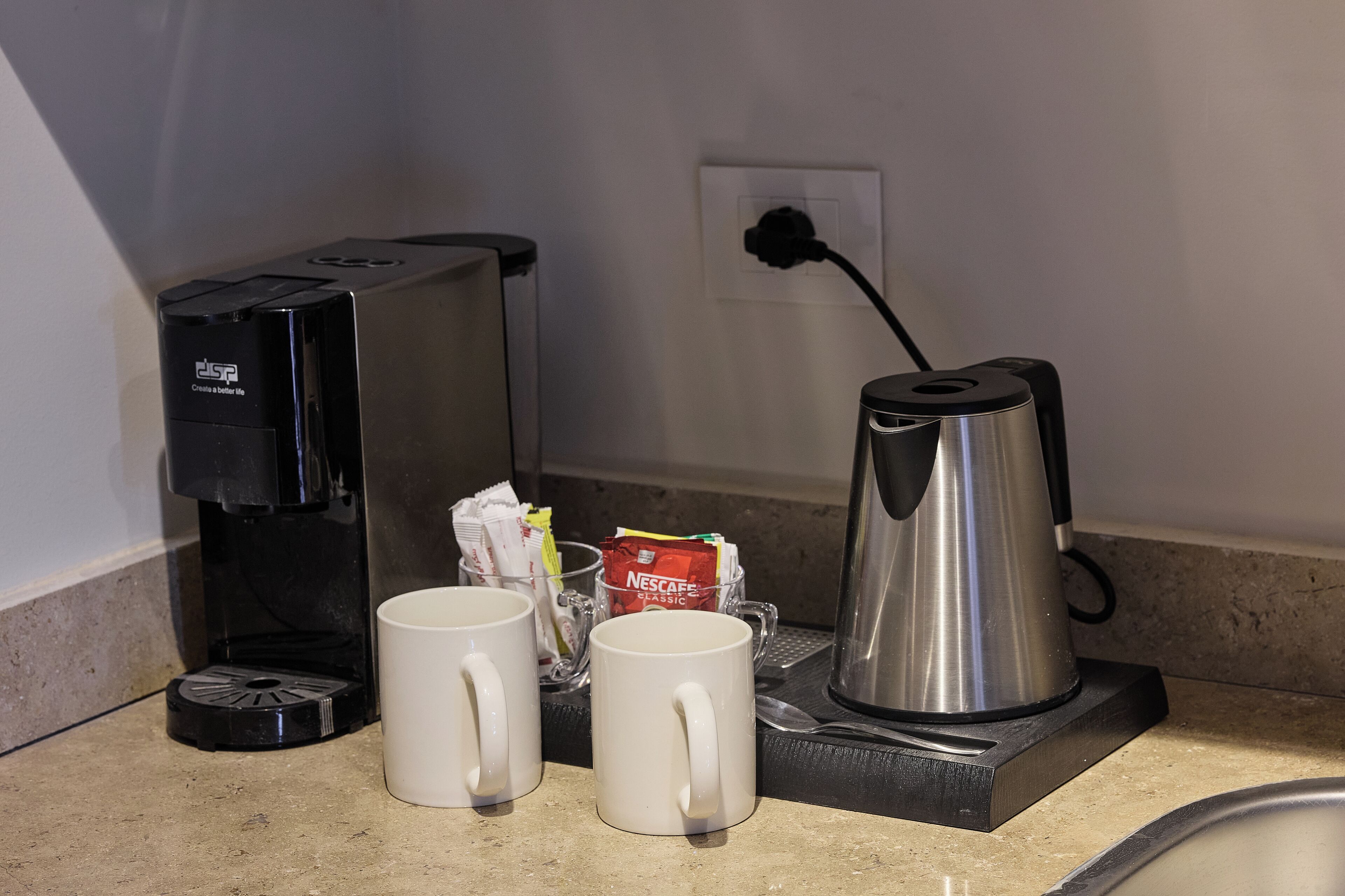 A coffee maker, two white mugs, a Nescafé Classic coffee pod, and a stainless steel electric kettle on a countertop.