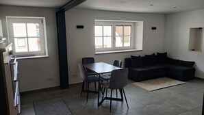 A modern living room with a dining area, featuring a black sofa, a dining table with chairs, and two windows providing natural light. - Modern vacation apartment in Kehl, 60m2 comfort on one level (Weißenburg in Bayern)