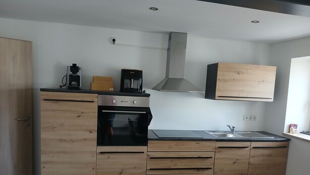 A modern kitchen with wooden cabinets, a built-in oven, a coffee maker, and a stainless steel sink. - Modern vacation apartment in Kehl, 60m2 comfort on one level (Weißenburg in Bayern)