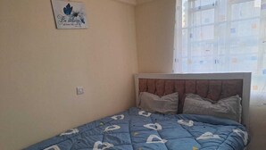 A bedroom with a bed, a wall-mounted picture, and a window with blinds. - Humble Homes 1 bedroom Thika Town CBD access to Netflix,Fridge,wifi (thika)
