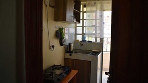 A compact kitchen with a gas stove, a sink, and a window with curtains. - Humble Homes 1 bedroom Thika Town CBD access to Netflix,Fridge,wifi (thika)