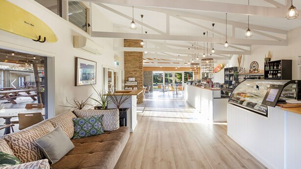 Un espacio interior amplio con suelo de madera, una zona de estar con sofá y cojines, una encimera de cocina y un comedor con mesas y sillas.