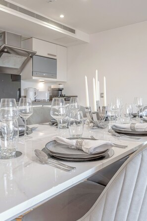 Un comedor moderno con una mesa de mármol blanco dispuesta para una comida, que incluye cubertería transparente, utensilios de plata y una decoración central.