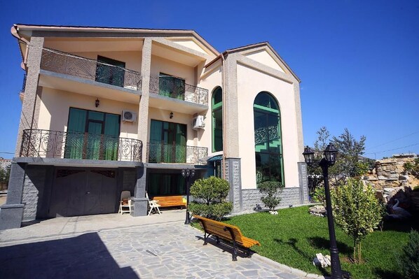 A two-story building with a balcony, a stone facade, and a small garden area with a bench and a lamp post.