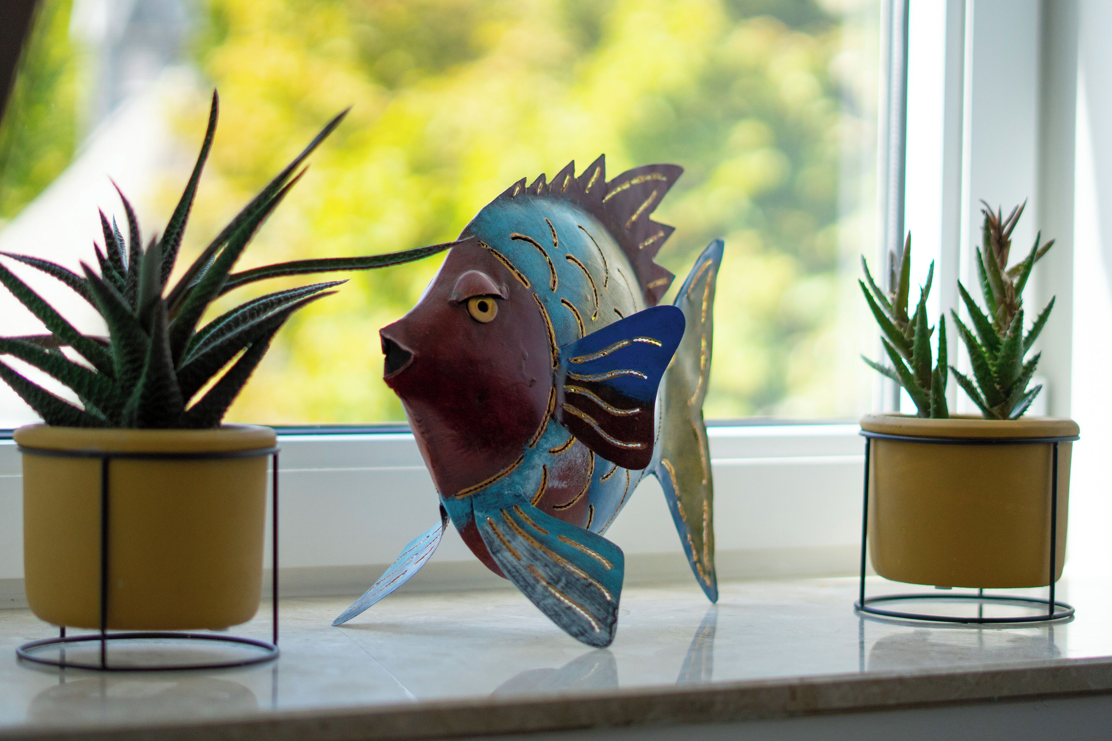 Une sculpture de poisson décorative est placée sur le rebord d’une fenêtre, avec deux plantes en pot de chaque côté.