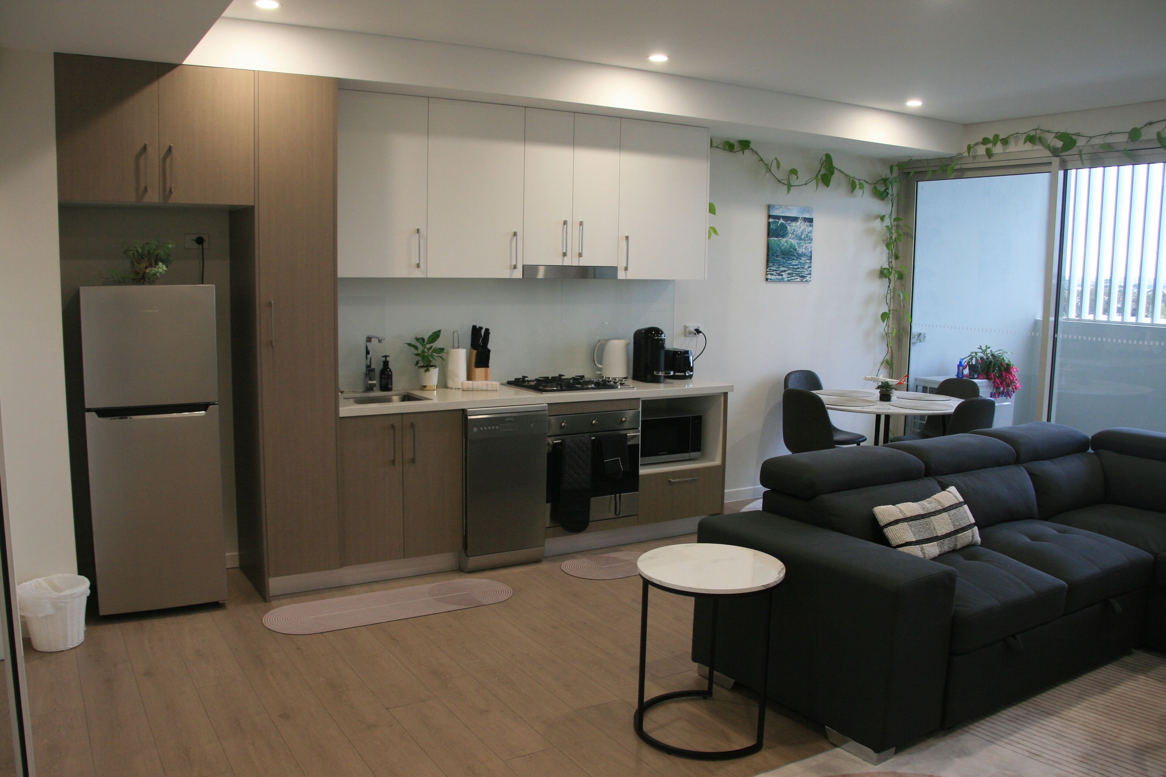 A modern kitchen with a refrigerator, stove, and dishwasher. There is a black sofa and a small round table in the living area.