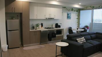 A modern kitchen with a refrigerator, stove, and dishwasher. There is a black sofa and a small round table in the living area.