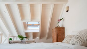 A bedroom with a high ceiling, a skylight, a bed with a white comforter, a wicker side table, and a wall-mounted lamp. - JOSY Atelier (Ferrel)