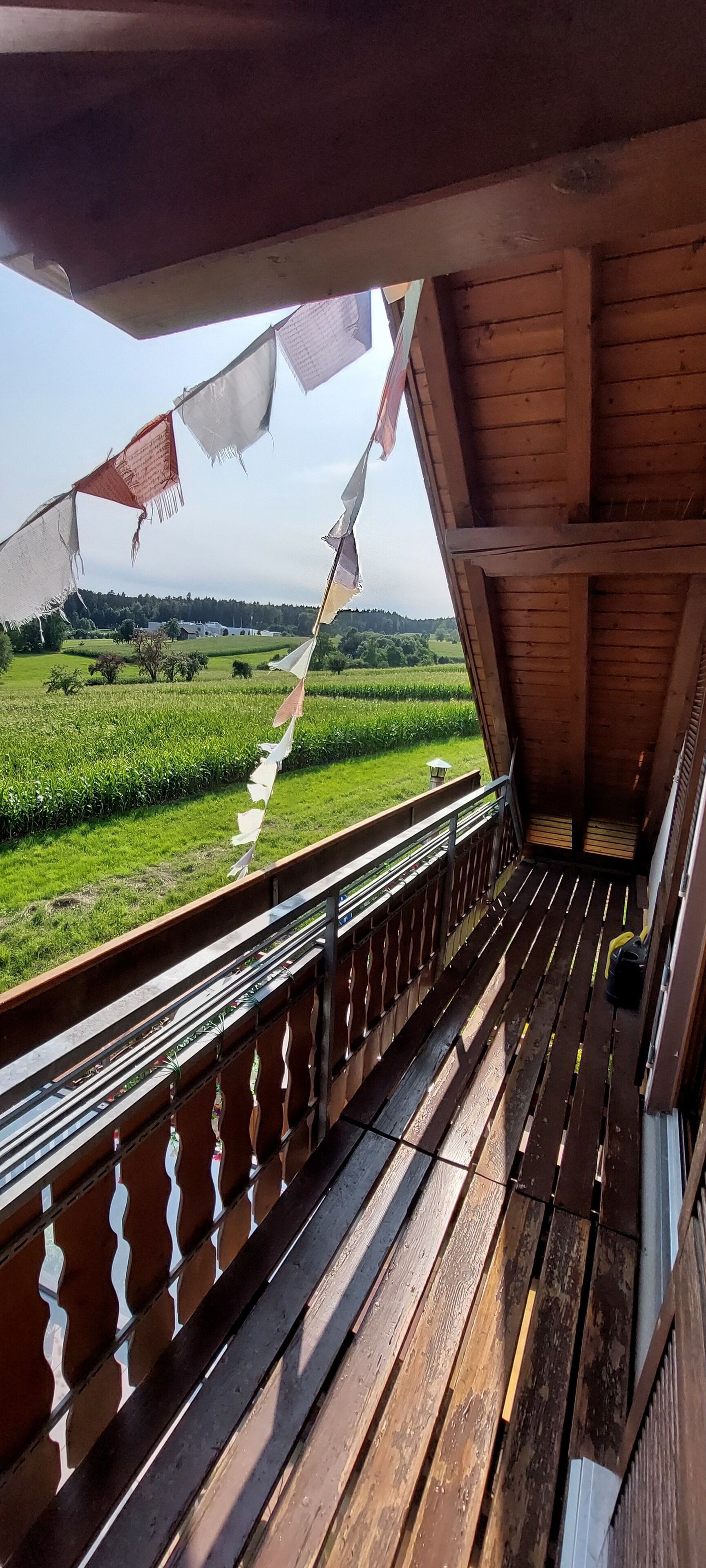 A wooden balcony with a view of green fields and a distant building.