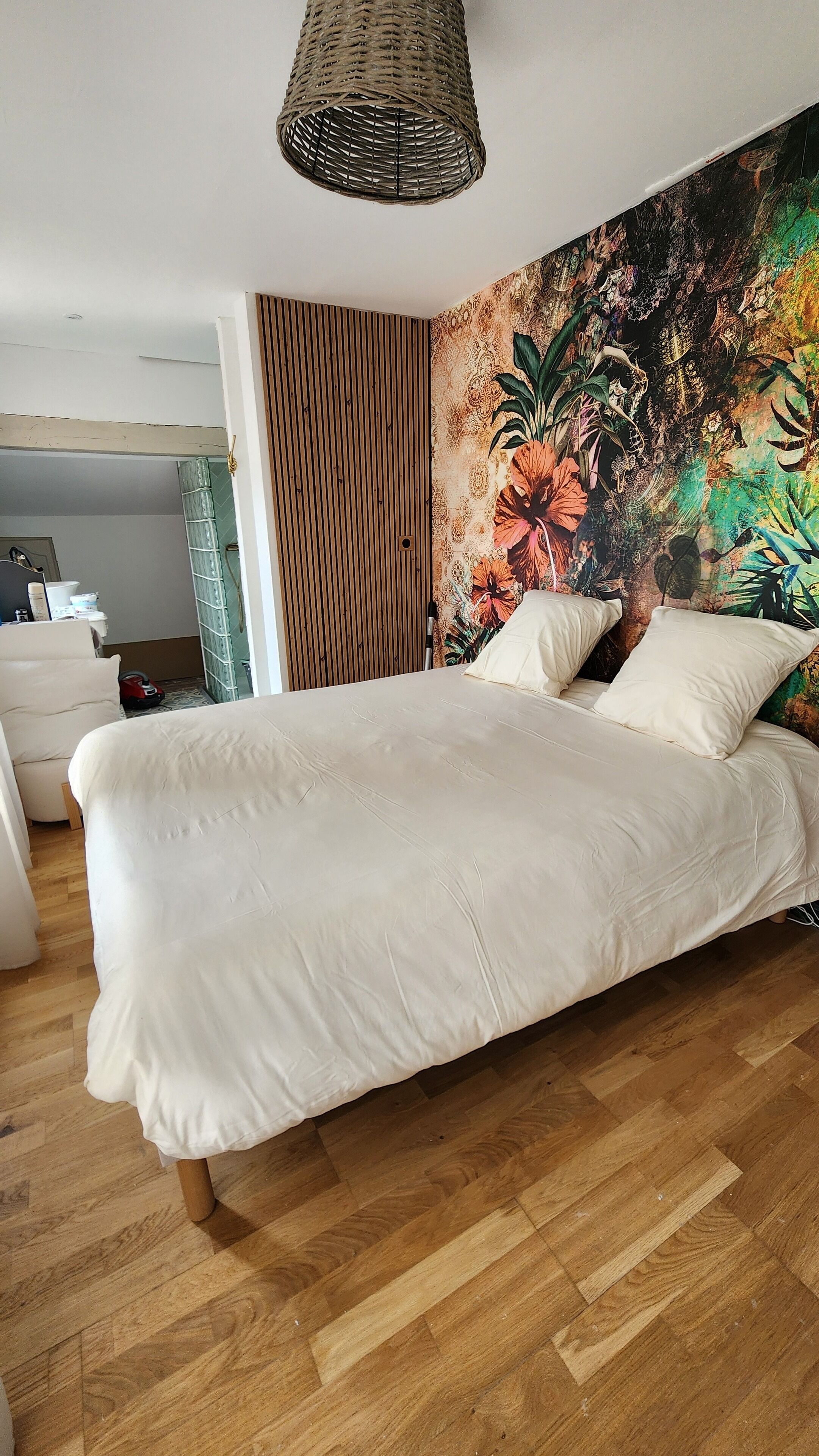A bedroom with a large bed, a wooden floor, a wall with a floral tapestry, and a hanging light fixture.