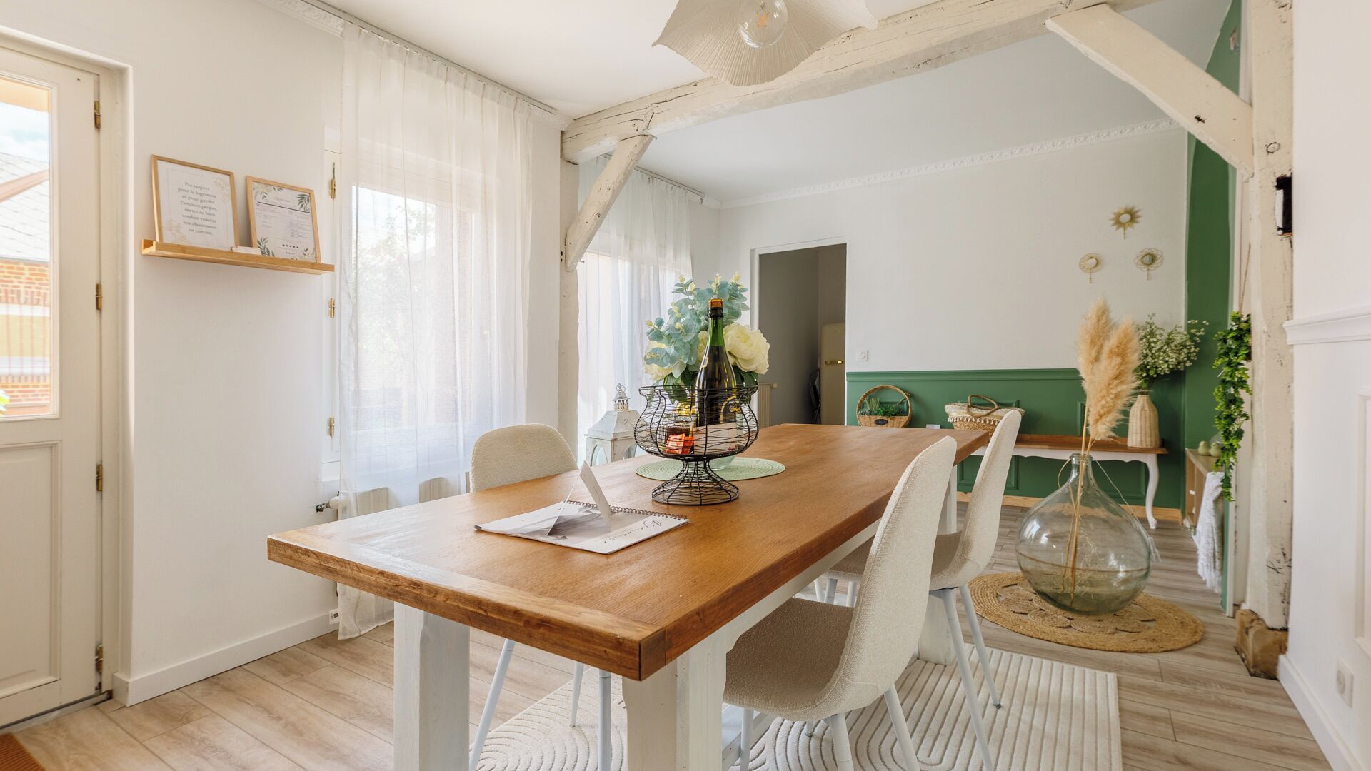 Un coin repas avec une table en bois, des chaises blanches, un vase avec des fleurs et une bouteille.