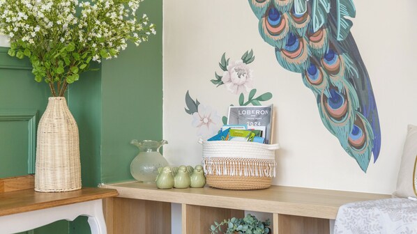 Ein Zimmer mit grüner Wand, einem Pfauen-Wandgemälde, einer Korbvase mit Blumen, einem Korb voller Bücher und einer kleinen Zimmerpflanze auf einem Regal.