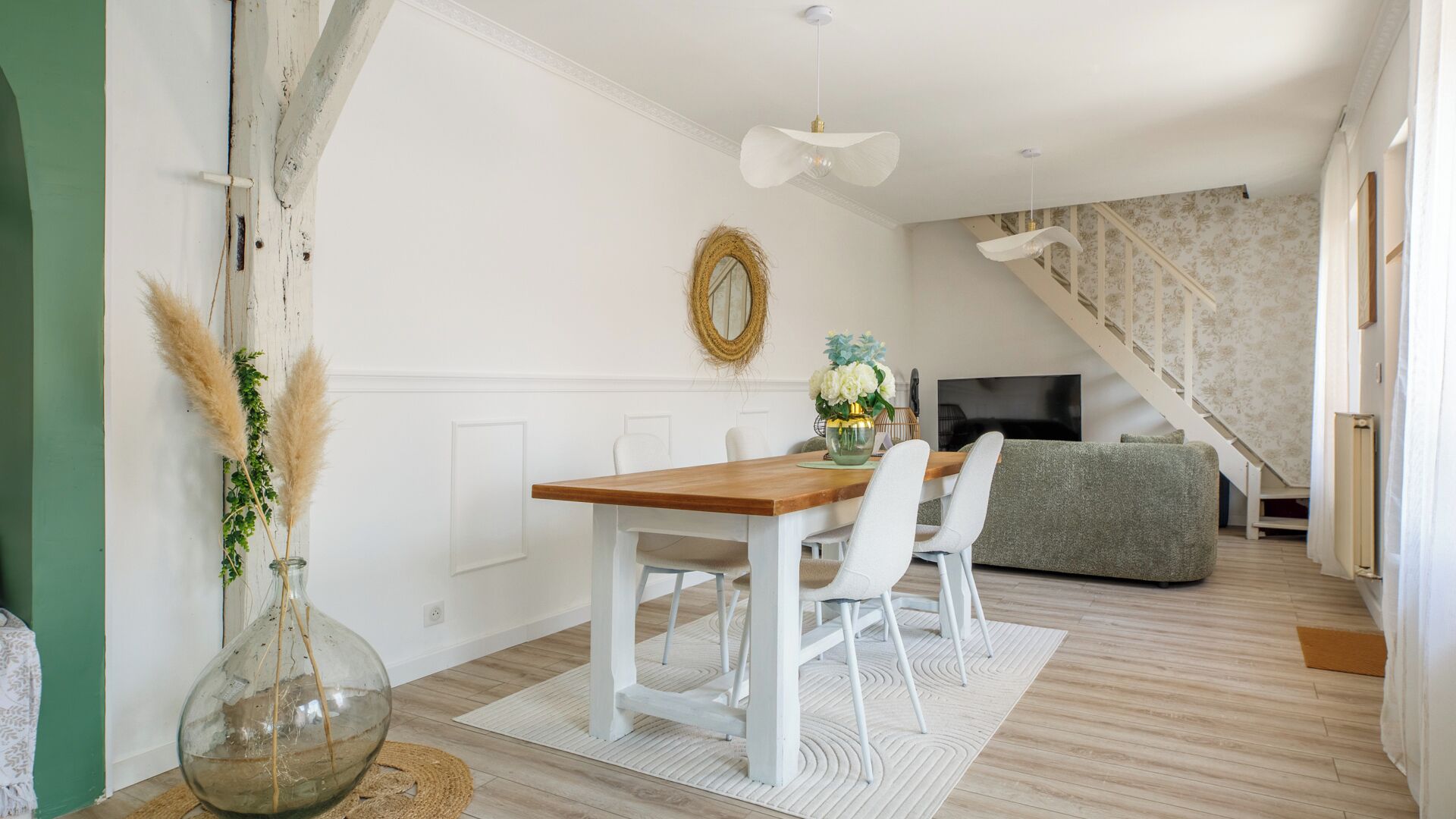 Ein modernes Wohnzimmer mit einem Holzesstisch, weißen Stühlen, einem grauen Sofa und einer Treppe mit ornamentalem Wandpapier.