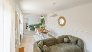 A cozy living room with a green sofa, a dining table with chairs, and a kitchen in the background.