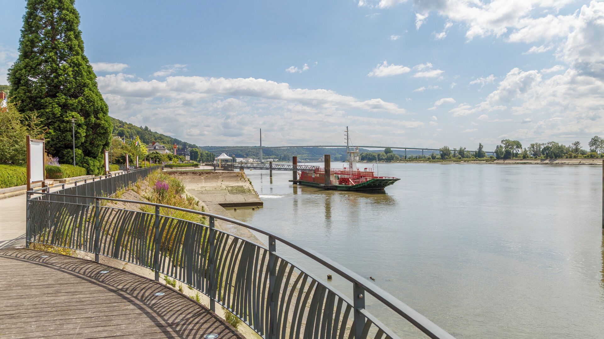 A riverside walkway with a metal railing, a dock with a boat, and a bridge in the background.