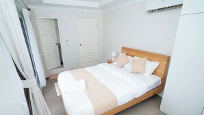 Een net opgemaakt bed met een houten frame, beddengoed in het wit en beige, en een nachtkastje met een lamp.