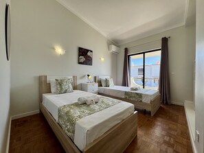 A bedroom with two beds, a nightstand, a wall-mounted air conditioner, and a large window with curtains. - ALBUFEIRA SUMMER VILLA 3 WITH POOL by HOMING (Albufeira)