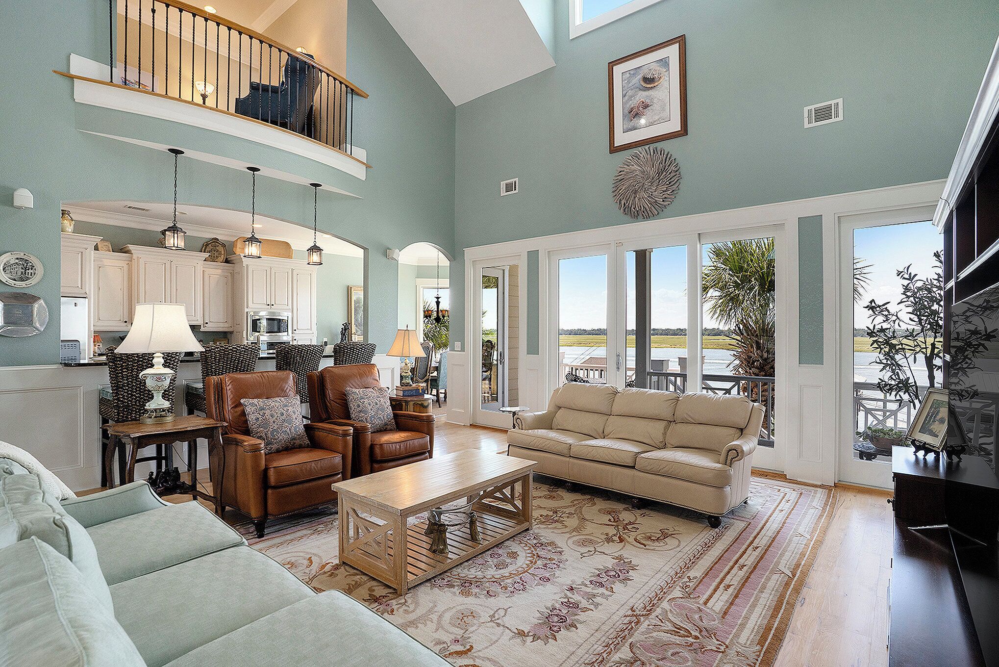 A spacious living area with a high ceiling, a dining area with white cabinets, and a large window offering a view of the outdoors.