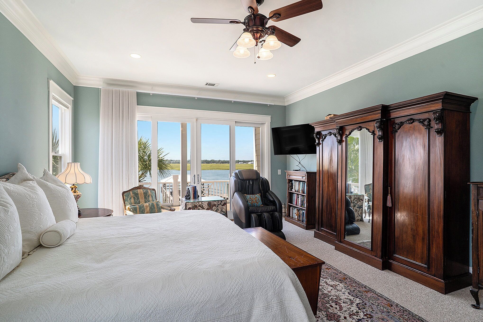 A bedroom with a large bed, a ceiling fan, a view of the outdoors, a massage chair, and a wooden cabinet.